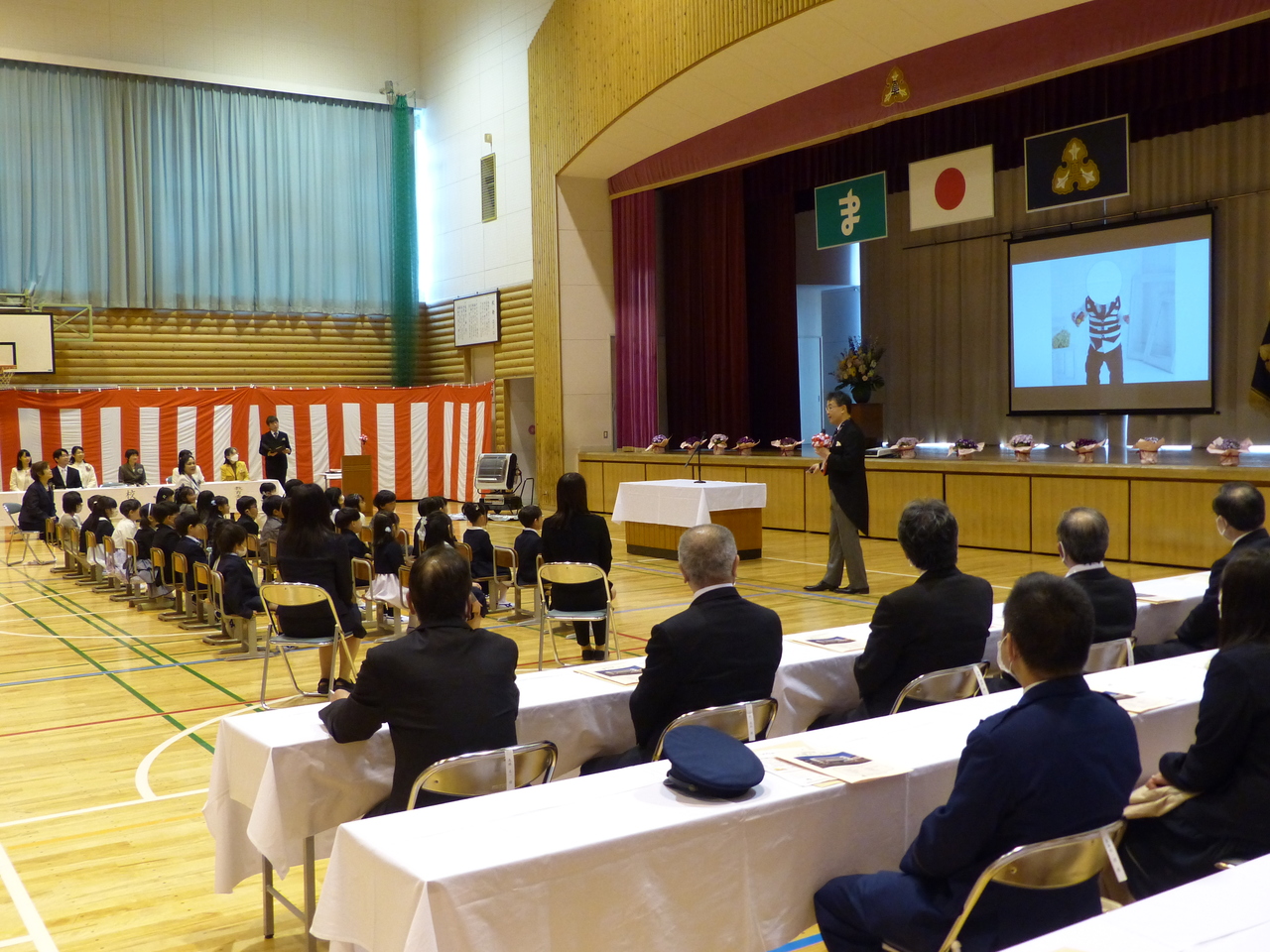 講壇の隣でマイクを手にもって話す燕尾服姿の男性の写真