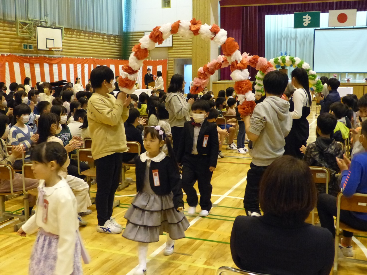 紙で出来た花輪のアーチをくぐって退場する児童たちの写真