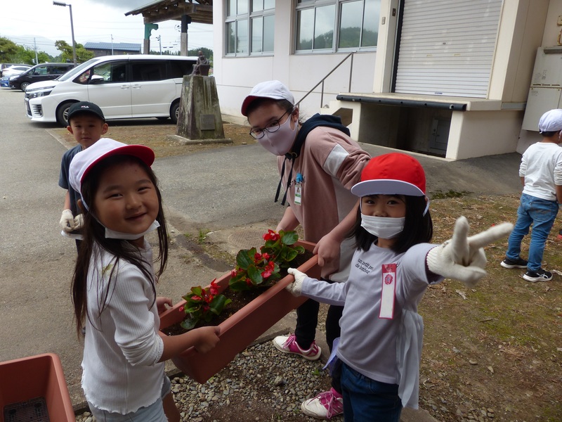 赤い花を植えたプランターを三人で持ち笑顔を向ける児童たちの写真
