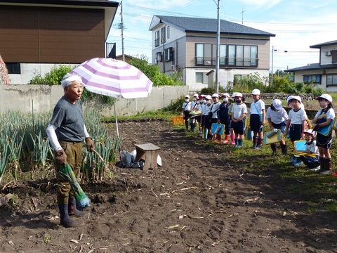 収穫した野菜を手に持つ作業着姿の男性と、畑の前に並ぶ児童たちの写真