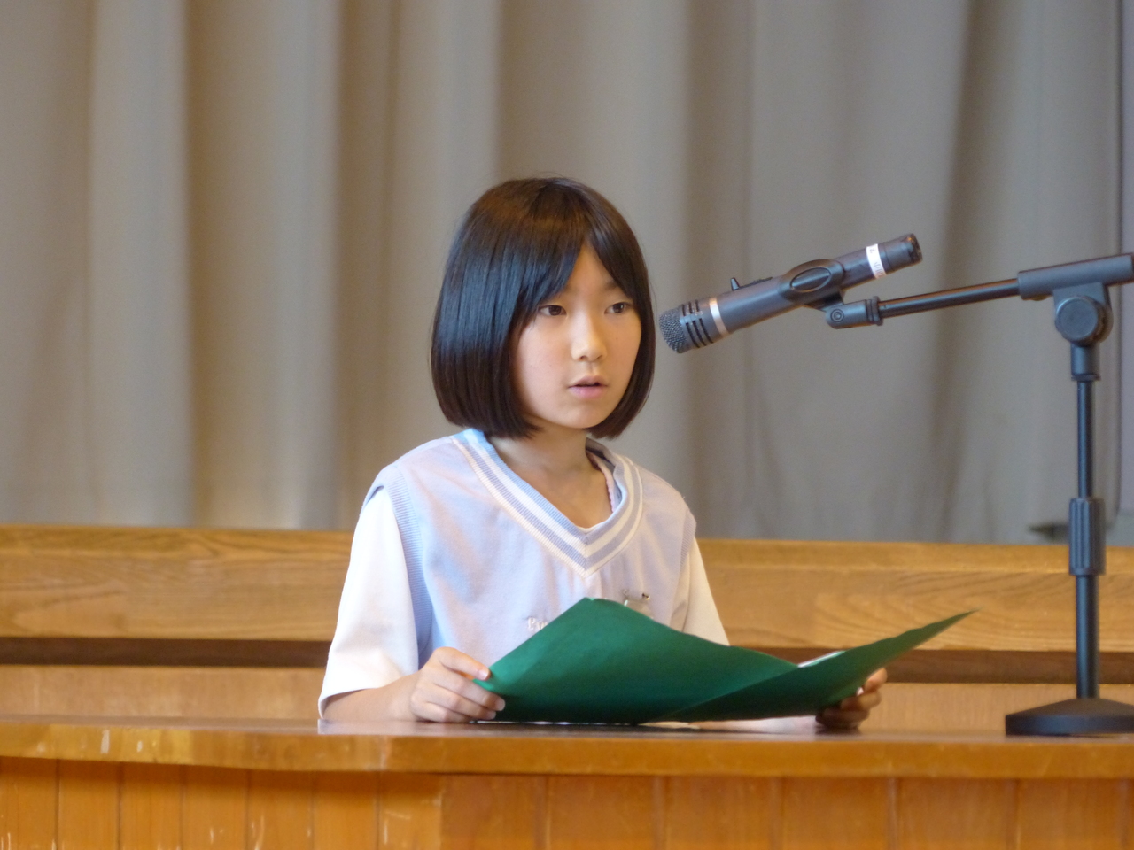 青緑色の冊子を持ちながらマイクの前で話す女子児童の写真