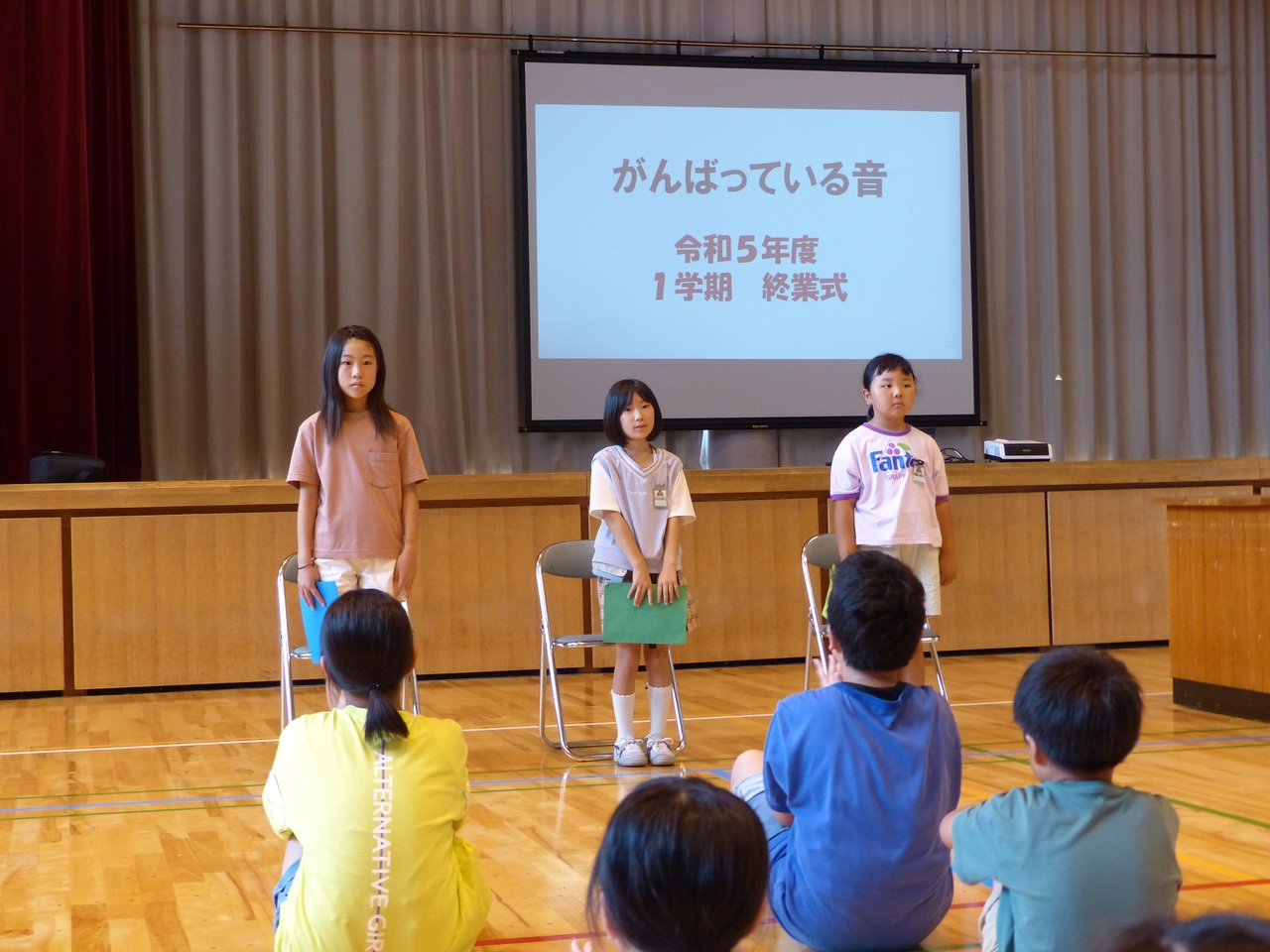 「がんばっている音 令和5年度1学期終業式」と書かれたモニターの前で起立する三人の女子児童の写真