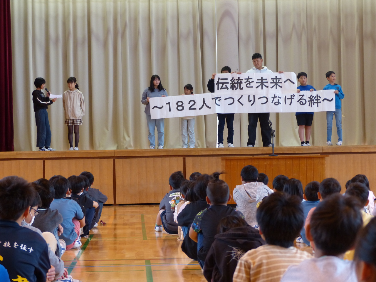 「伝統を未来へ～182人でつくりつなげる絆～」と書かれた大きな紙を6人がかりで手に持っている子供たちの写真