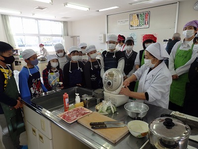 エプロンとキャップとマスクを身につけた児童たちが、炊飯器を扱う先生の様子を熱心に観ている写真