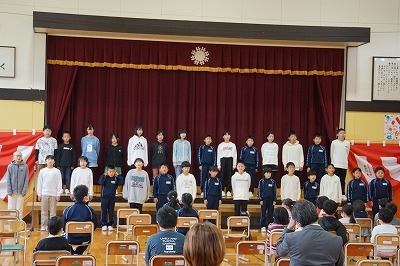 体育館の舞台上とその手前にいっぱいに広がって歌う「童唄クラブ」の児童たちの写真